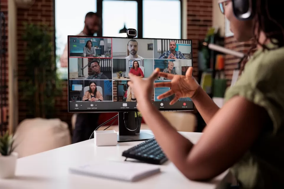 Person wearing headphones sitting at desk with monitor showing other people on Zoom meeting.